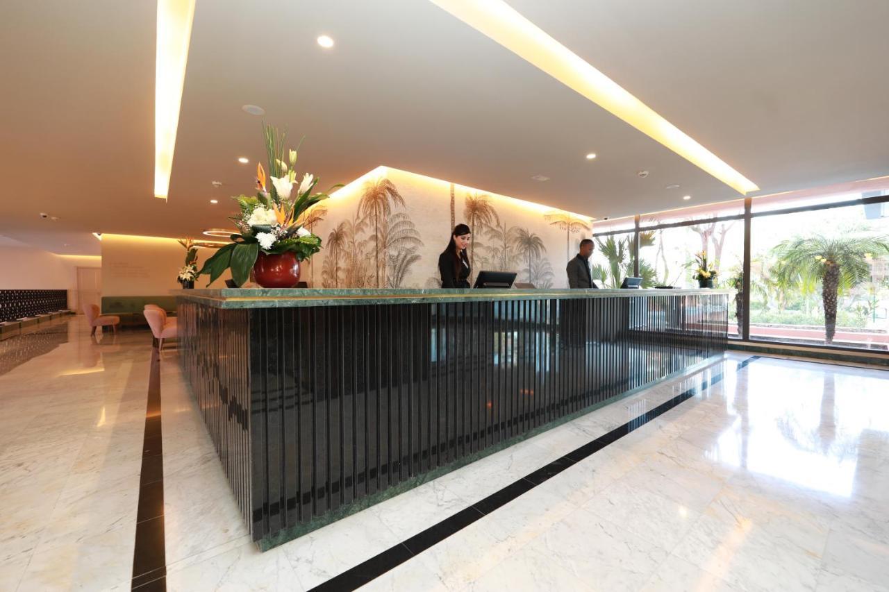Hotel Kenzi Rose Garden Marrakesh Esterno foto The image shows a modern reception area, likely for a hotel or office building. There is a stylish front desk made of dark materials with a smooth surface. Behind the desk, two staff members are engaged in their work, possibly checking in guests or a